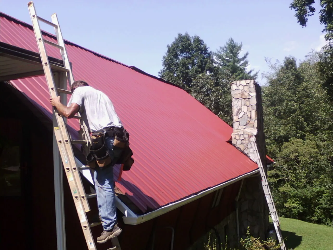 Roofer on a ladder beside a red metal roof and stone chimney — Soffit & Fascia Repair by Roofing Brothers in Waskom